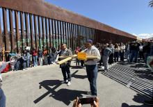 A participant in a symbolic Via Crucis held Saturday in Nogales, Mexico, to honor migrants uses a smartphone (at bottom) to take a picture of the group that includes a man holding a wooden cross next to the border wall with Arizona. The orange sign reads,"We are the body of Christ." (Anita Snow)
