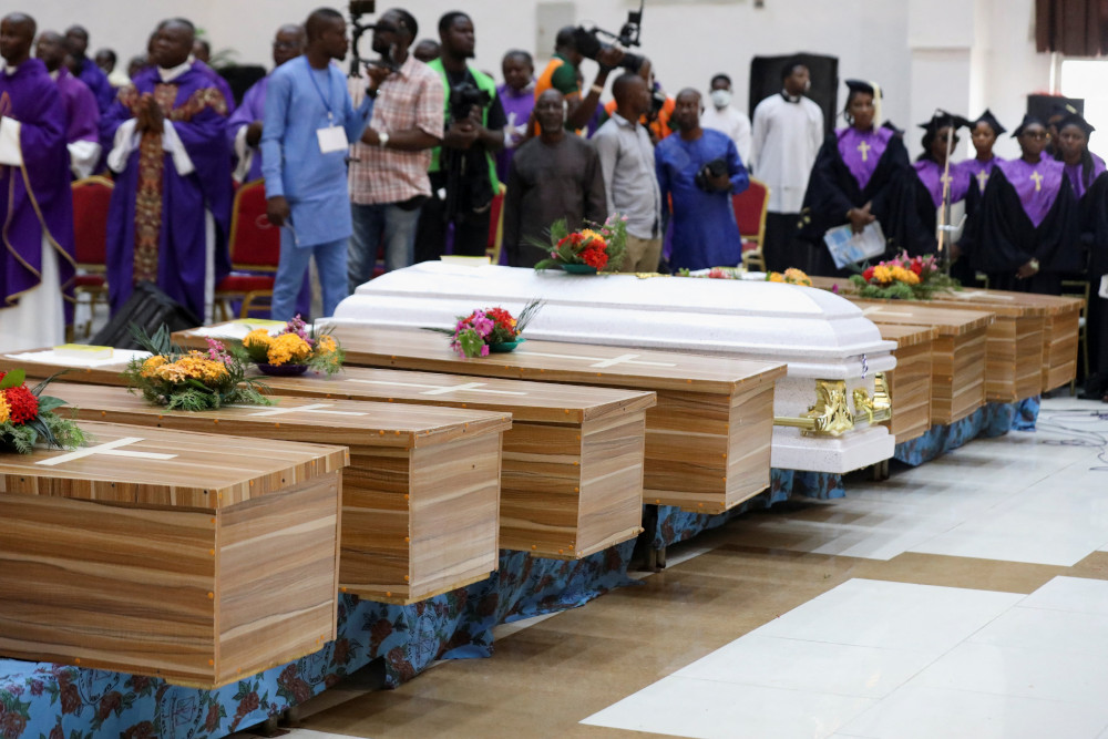 A row of caskets have flowers placed on them. They are surrounded by people in various church garments and other people with news cameras.