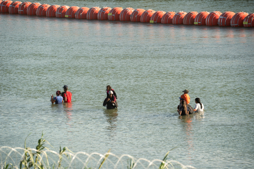 People stand in the middle of a river. Some have others on their shoulders. One part of the river is blocked by large orange buoys spaced so close as to be a wall and the other side is blocked by concertina wire.