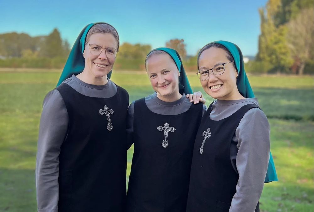 Three women stand together in a field. They are wearing back and gray dress with a cross on the front and teal veils.
