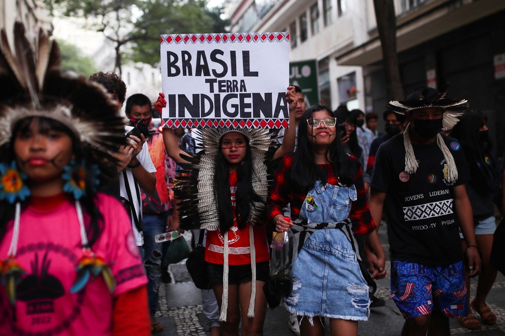 Brazilian bishops, Land disputes. People wearing indigenous garb and ornamentation march in street; one person holds sign reading 'Brazil is Indigenous Land'.