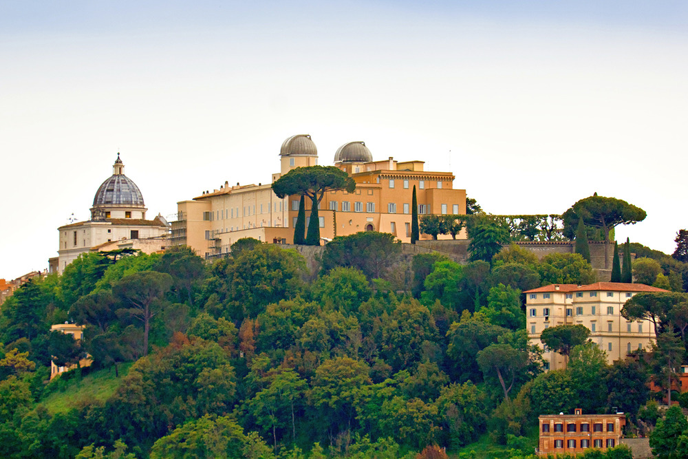 Specola Vaticano A cluster of large, romanesque buildings sits stop a verdant hill