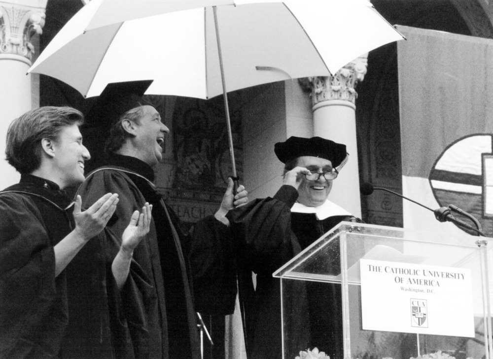 Bob Newhart, Catholic University of America commencement, 1997. Black and white photo of the three gathered under umbrella in front of lectern.