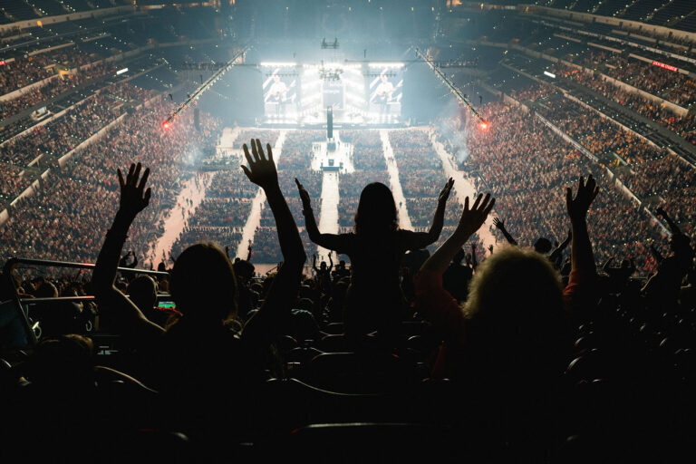 National Eucharistic Congress, July 20, 2024. Back view of people in nosebleed seats raising hands; light emanates from distant stage.