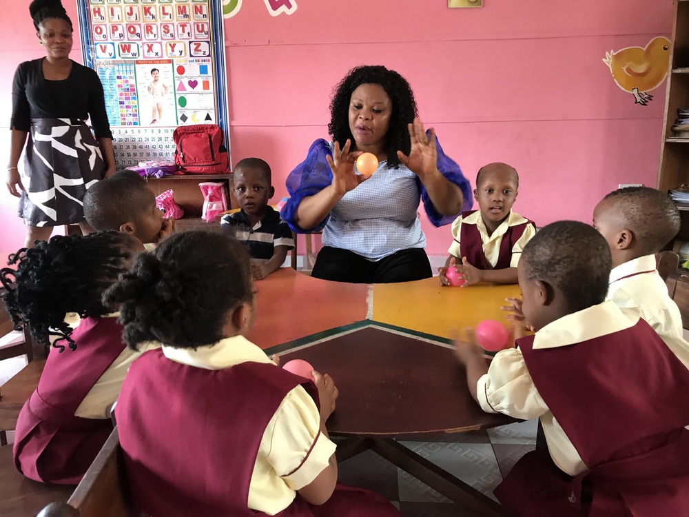 Catholic Montessori school in Uyo, Nigeria, March 9, 2021. Teacher sits at table with students, holding a yellow sphere.