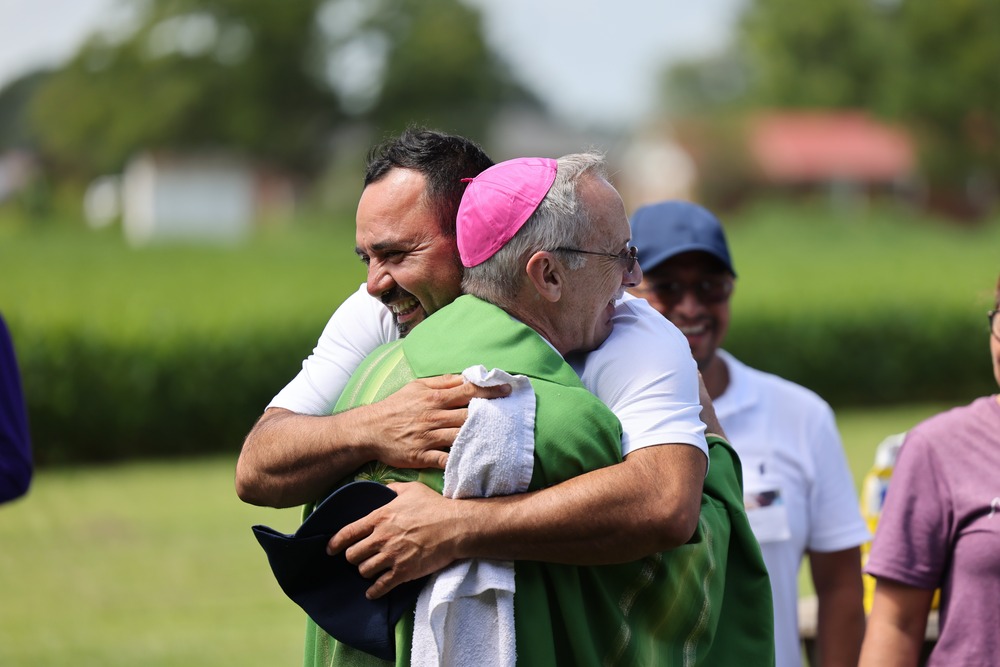 Bishop Luis R. Zamara of Raleigh at the XIII Encuentro Catolico de Campesinos, Aug. 4, 2024. The bishop, vested in green, hugs a smiling man; in the background is a field of tall green stalks, possible corn.