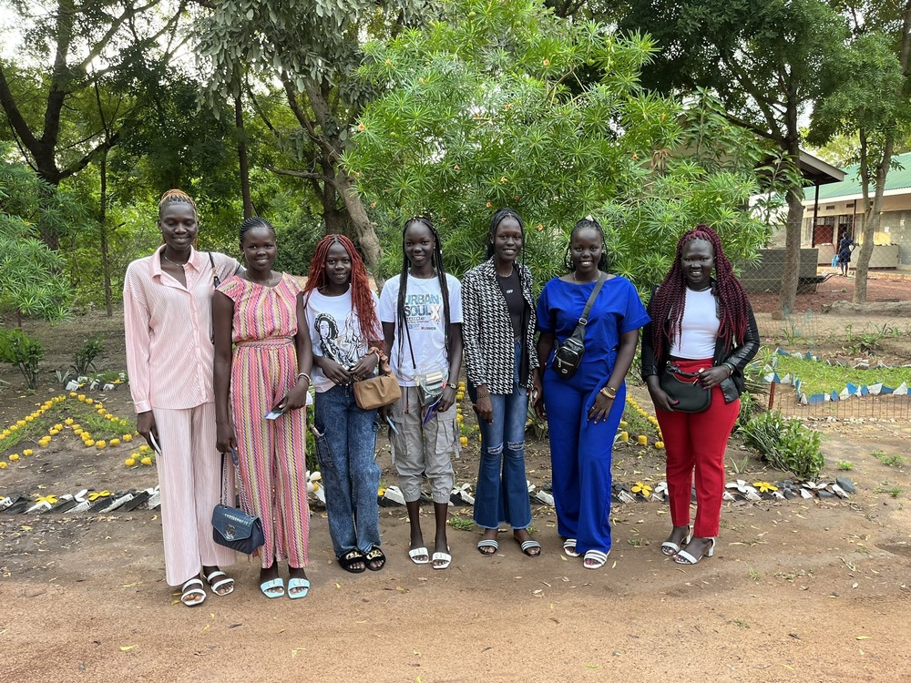 Graduates of Loreto Rumbek in South Sudan. Seven young women stand in row. In the background are trees, garden beds, and buildings.