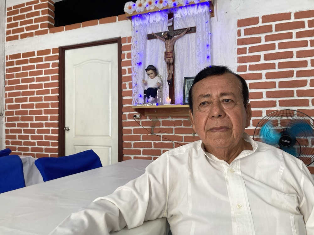Daniel Hernández Rabanales, Oct. 9, 2024, Huixtla, Mexico. Rabanales sits at a table, behind him is a crucifix and the El Nino Dios dressed as a migrant in a small wall shrine.