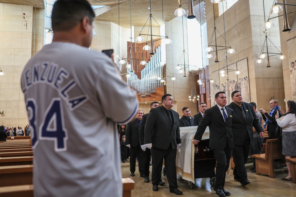 Funeral of Fernando Valenzuela, Cathedral of Our Lady of Angels, Los Angeles, Nov. 2024. In foreground is back of fan wearing Valenzuela jersey, centered are pall bearers carrying casket through Cathedral aisle.