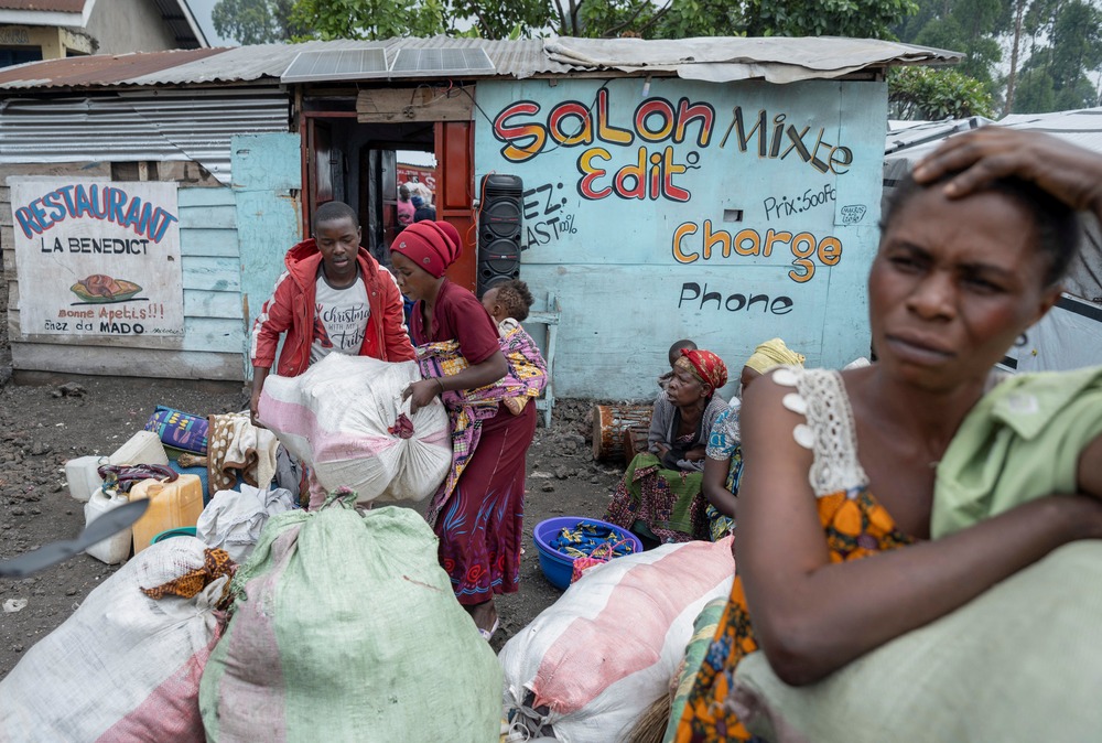 Displaced Congolese, North Kivu, Congo, Feb. 3, 2025. A woman is foregrounded leaning on sacks with hand on head, in background is a small storefront and and other people, including a woman with a baby on her back.