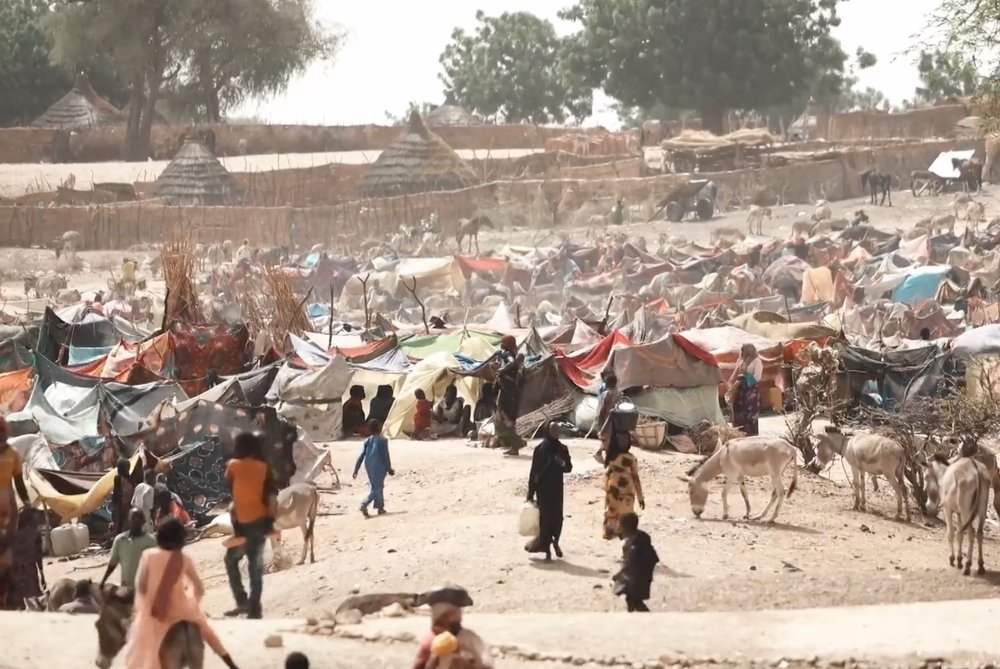 View of many people walking and sitting among tents in dusty area. 