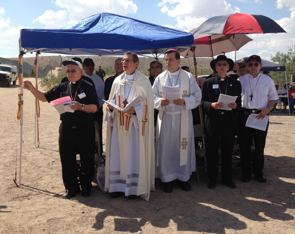 Archbishop Gustavo Garcia-Siller of San Antonio, Pearsall, Tx. The archbishop wears white vestments and reads from book, others assembled around under tarpaulin tent and umbrellas.
