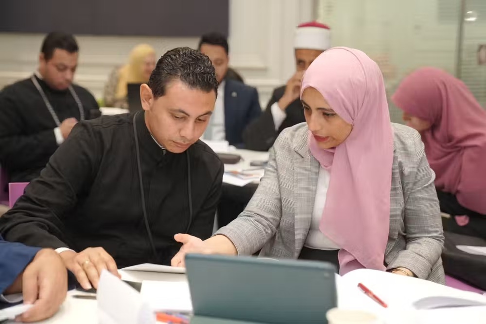 Climate change and religion workshop, Egypt, 2023. Benadi wearing business garb and pink hijab sits at a table with clergyman explaining concepts.