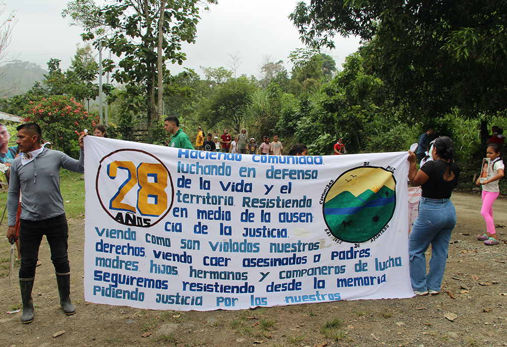 Miembros de la comunidad sostienen un cartel durante su procesión hacia las tumbas de los miembros asesinados en 2024. (Foto: Tracy L. Barnett) Miembros de la comunidad sostienen un cartel durante su procesión hacia las tumbas de los miembros asesinados en 2024. (Foto: Tracy L. Barnett)