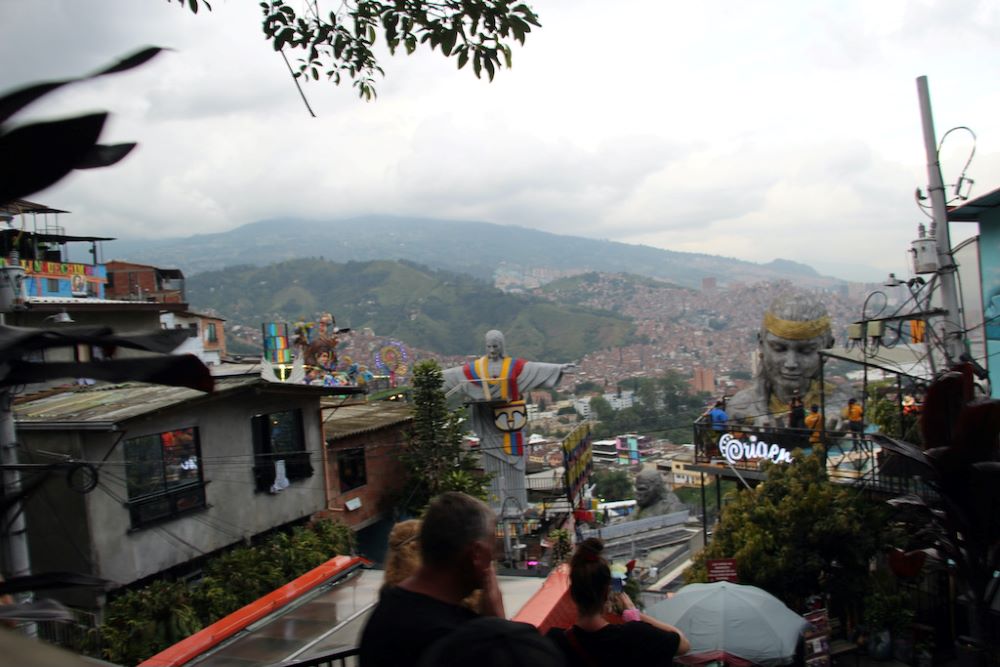 View includes flashing neon and tin-roofed homes stacked up the hillside. A tricolor-draped Christ, a massive gorilla and a concrete woman's head, an icon representing a local craft beer called “Origen,”overlook the scene.  View includes flashing neon and tin-roofed homes stacked up the hillside. A tricolor-draped Christ, a massive gorilla and a concrete woman's head, an icon representing a local craft beer called “Origen,”overlook the scene.