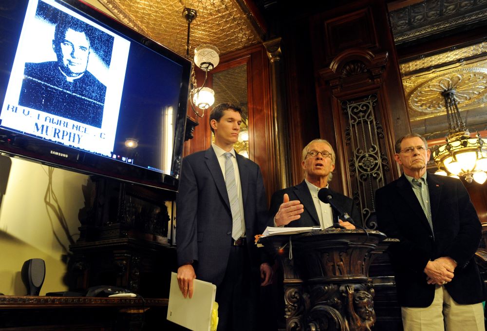 Three men at a news conference, with image of Fr. Lawrence Murphy on monitor