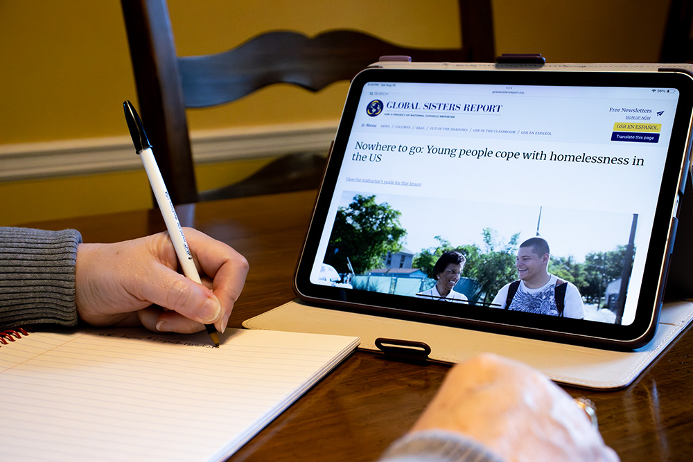 A screen on a tablet shows Global Sisters Report while a person takes notes in a notebook are visible. (GSR photo illustration/Teresa Malcolm)