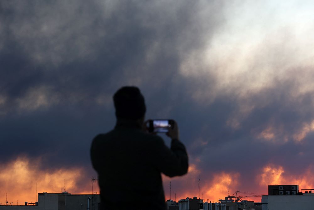 A person uses a phone as smoke rises following an explosion in Tehran, Iran, March 3. Israel and the U.S. launched strikes on Iran Feb. 28. (OSV News/West Asia News Agency via Reuters/Majid Asgaripour)