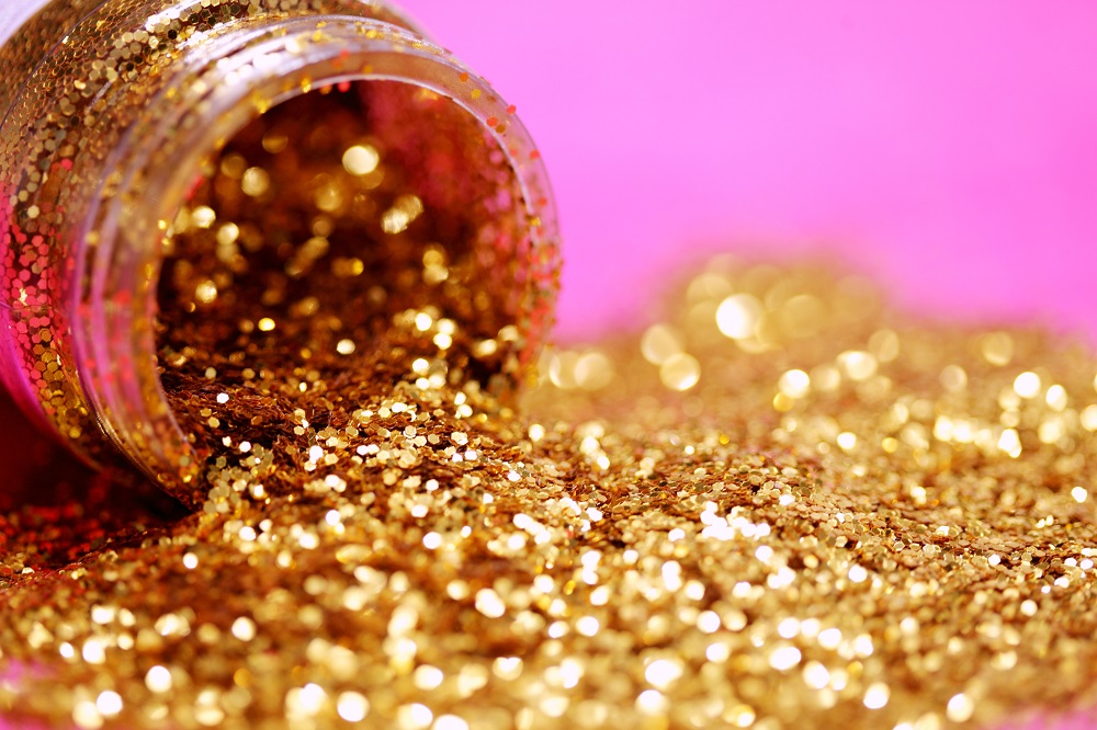 (Photo by Sharon McCutcheon on Unsplash) Image: A bottle of gold glitter lies on its side, glitter in a pile on a pink background.