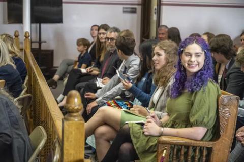 Montanan youth in court against state due to fossil fuel policies. Rows of young people sit in courtroom benches; one smile in direction of camera.