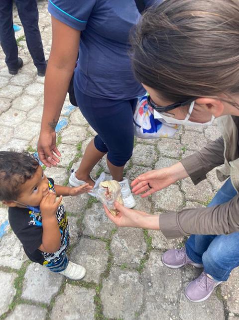 La hermana Dondé ofrece una alimento a un niño migrante que es llevado de la mano por su madre.
